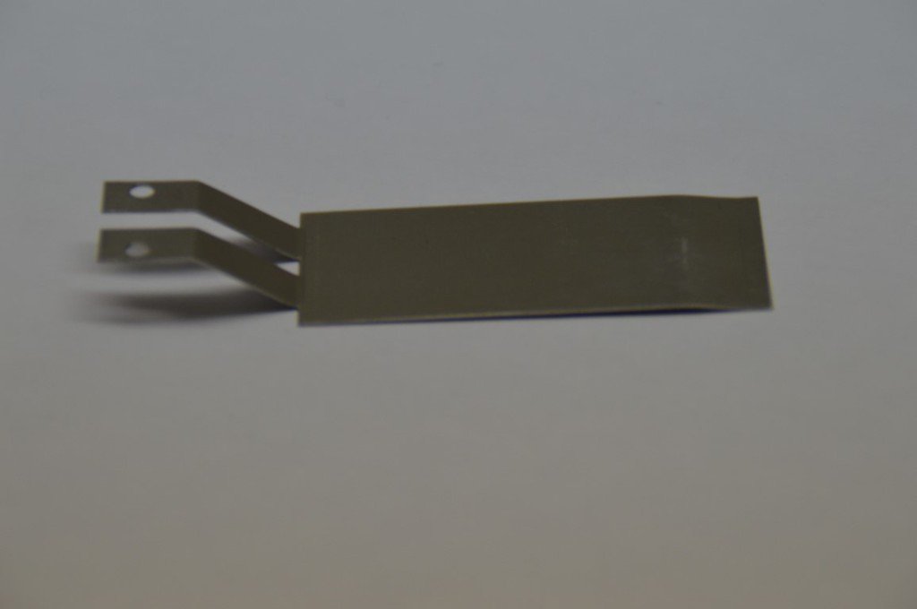 Dünnes Metallteil mit zwei gelochten Anschlusszungen und gebogenem Schaft – gefertigt als elektrischer Kontakt oder Federbauteil